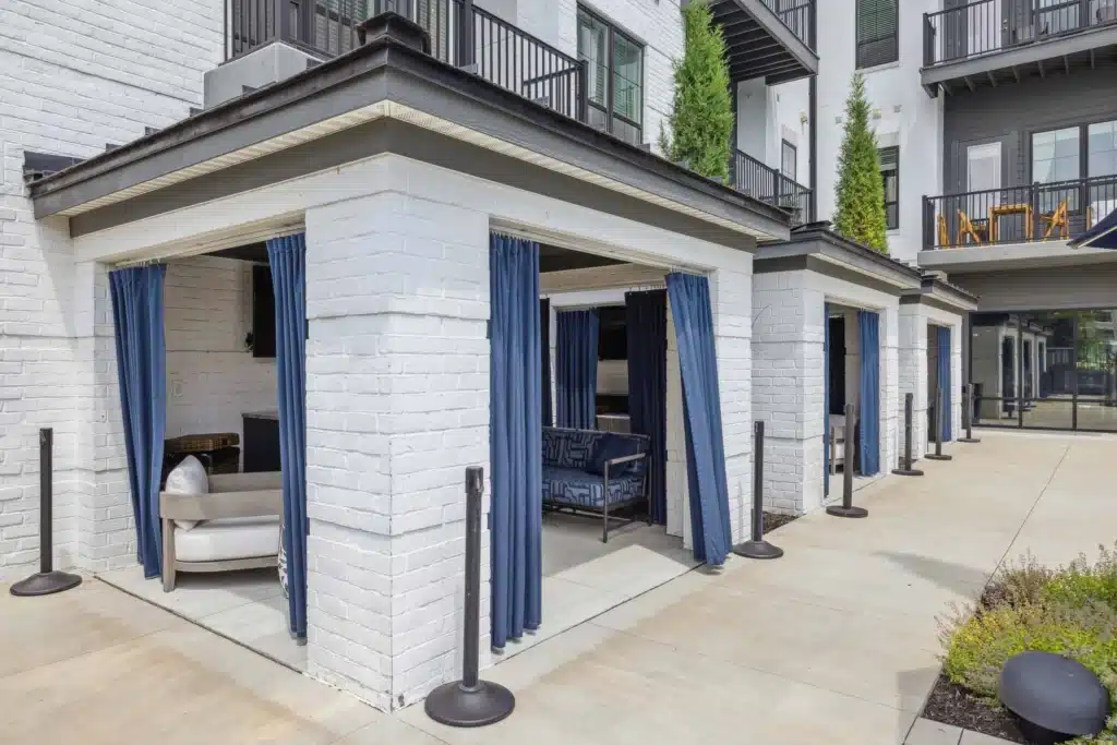 Outdoor cabanas with white brick walls, blue curtains, and seating areas are lined up outside a modern apartment building.