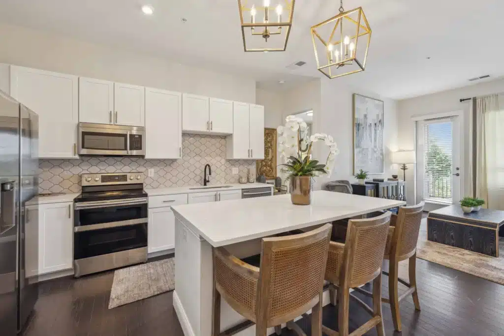 Modern kitchen with white cabinets, stainless steel appliances, an island with four wicker chairs, pendant lights, and a view of the living area and balcony.