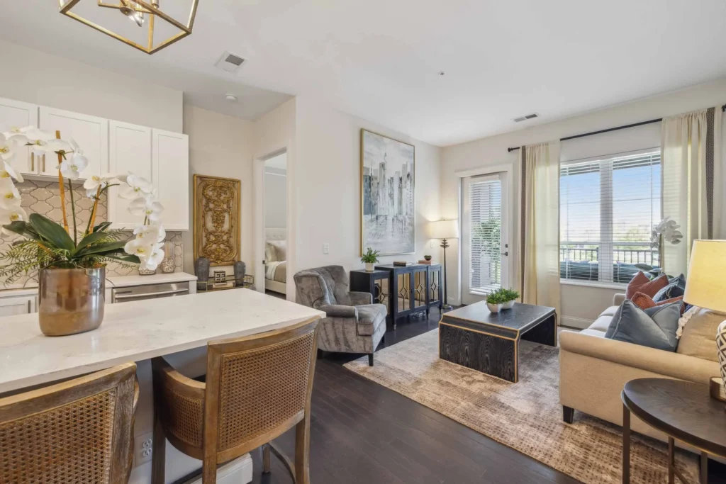 Modern living room with neutral decor, a sofa, two chairs, coffee table, and kitchen island with barstools; large windows and balcony door let in natural light.