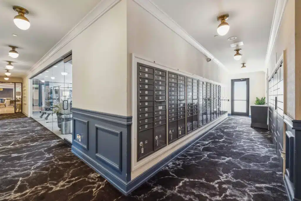 A hallway with rows of black mailboxes mounted on the wall; glass doors reveal a gym on the left, and patterned carpet covers the floor.