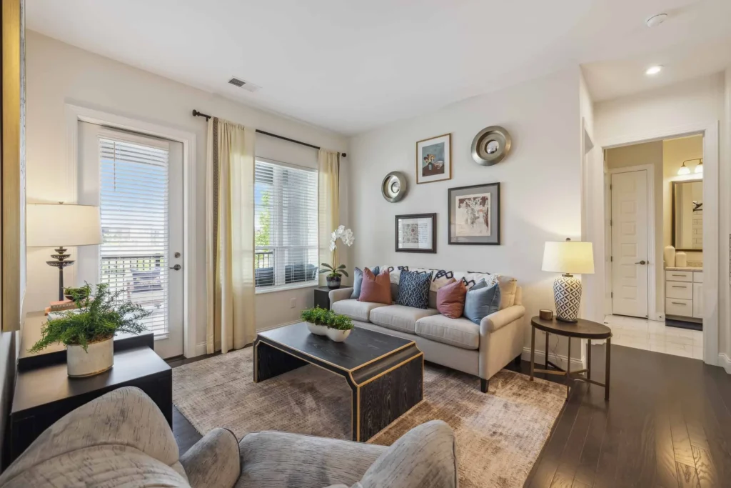 Modern living room with a beige sofa, armchair, black coffee table, decorative pillows, and large windows with sheer curtains; adjacent to an open hallway and bathroom.