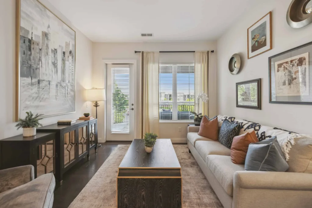 A modern living room with a beige sofa, wooden coffee table, large wall art, sideboard, and framed pictures. Light streams in through windows and a glass door with light curtains.