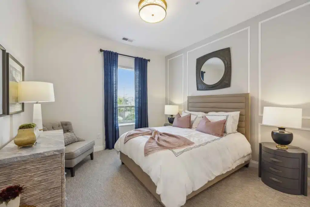 Modern bedroom with a bed, two bedside tables with lamps, a dresser, a gray chair, blue curtains, and a large round mirror above the headboard.