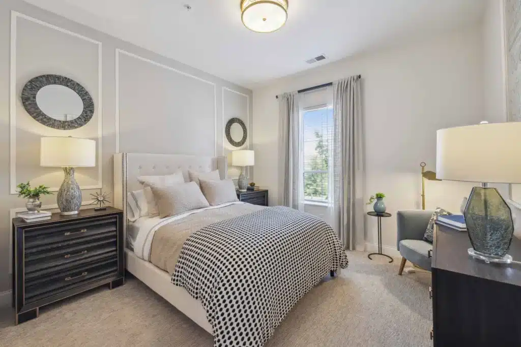 Modern bedroom with a neatly made bed, two nightstands with lamps, a desk and chair, large window with curtains, and decorative mirrors on the wall.