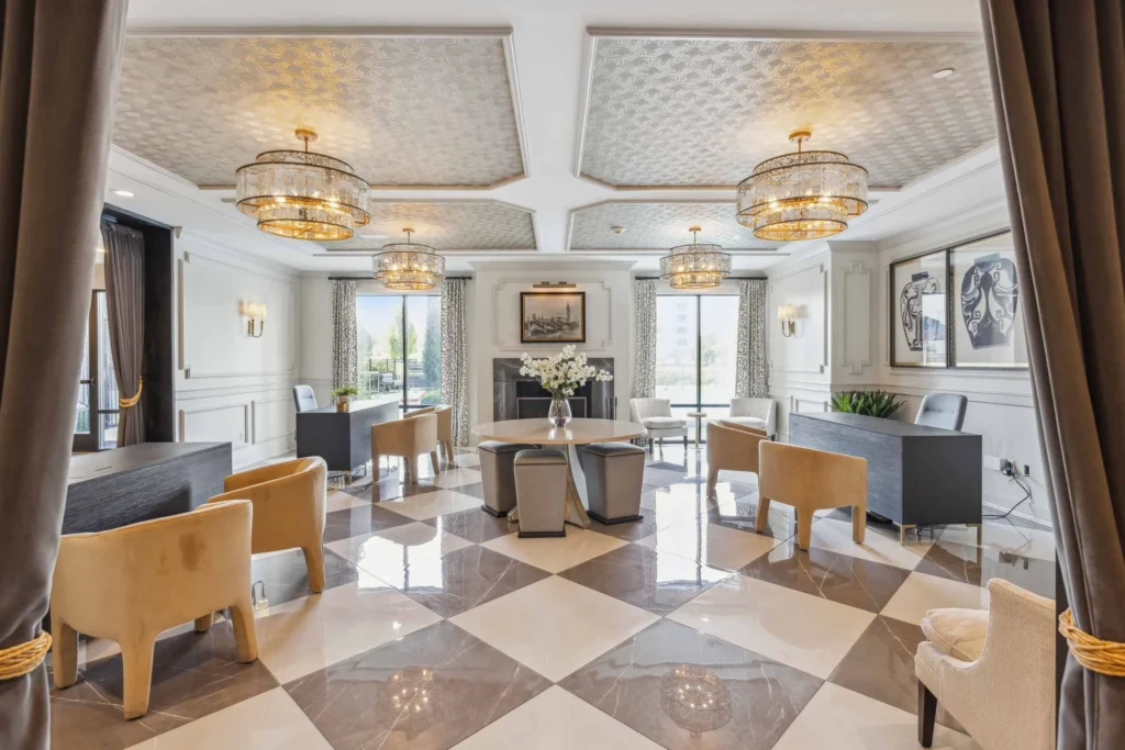 Elegant office lobby with checkered tile floors, modern chandeliers, multiple desks with chairs, large windows, and framed artwork on white paneled walls.