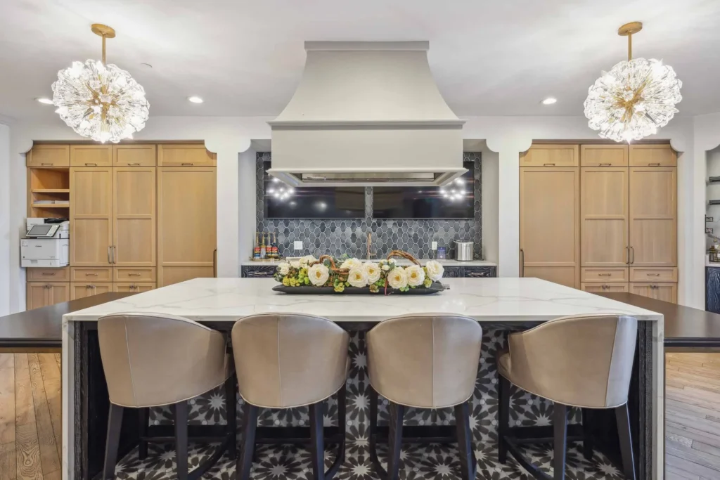 Modern kitchen with a large island, four beige barstools, light wood cabinets, geometric tile backsplash, and two decorative pendant lights. A floral arrangement sits on the island.