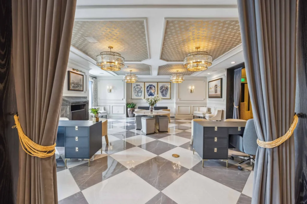 A modern, elegant office lobby with gray curtains, checkered tile flooring, chandeliers, and two desks facing each other, with artwork and plants in the background.