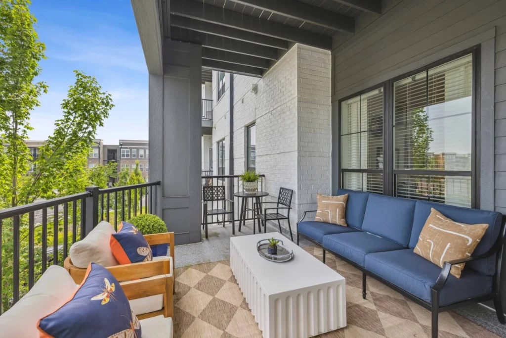 A modern apartment balcony with outdoor seating, a white coffee table, blue cushions, accent pillows, and a small dining set, overlooking a residential area.