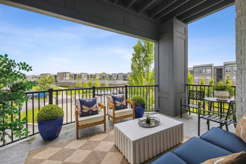 Covered balcony with outdoor seating, potted plants, and a coffee table overlooks a landscaped apartment complex under a blue sky.