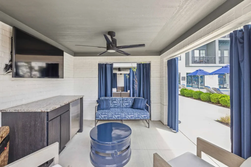 A covered outdoor lounge area with a blue patterned sofa, wall-mounted TV, ceiling fan, small counter with mini fridge, and blue curtains opening to a poolside view.
