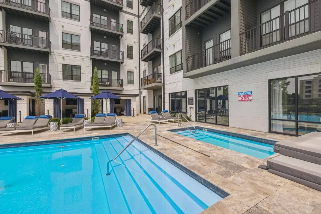 Outdoor apartment pool area with lounge chairs, blue umbrellas, hot tub, and modern multi-story building in the background.