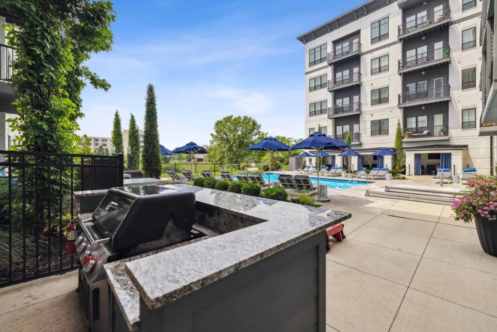 Outdoor pool area with lounge chairs, blue umbrellas, and a barbecue grill station next to a modern apartment building on a clear day.