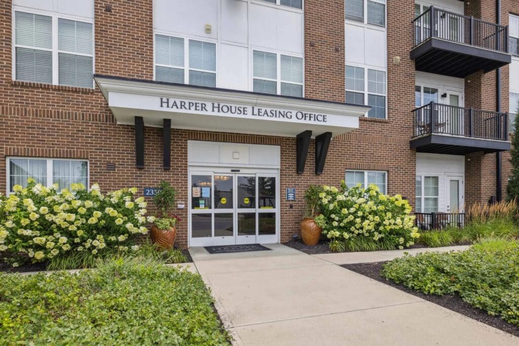 Entrance to Harper House Leasing Office, featuring glass doors, potted plants, and surrounding greenery in front of a brick apartment building.