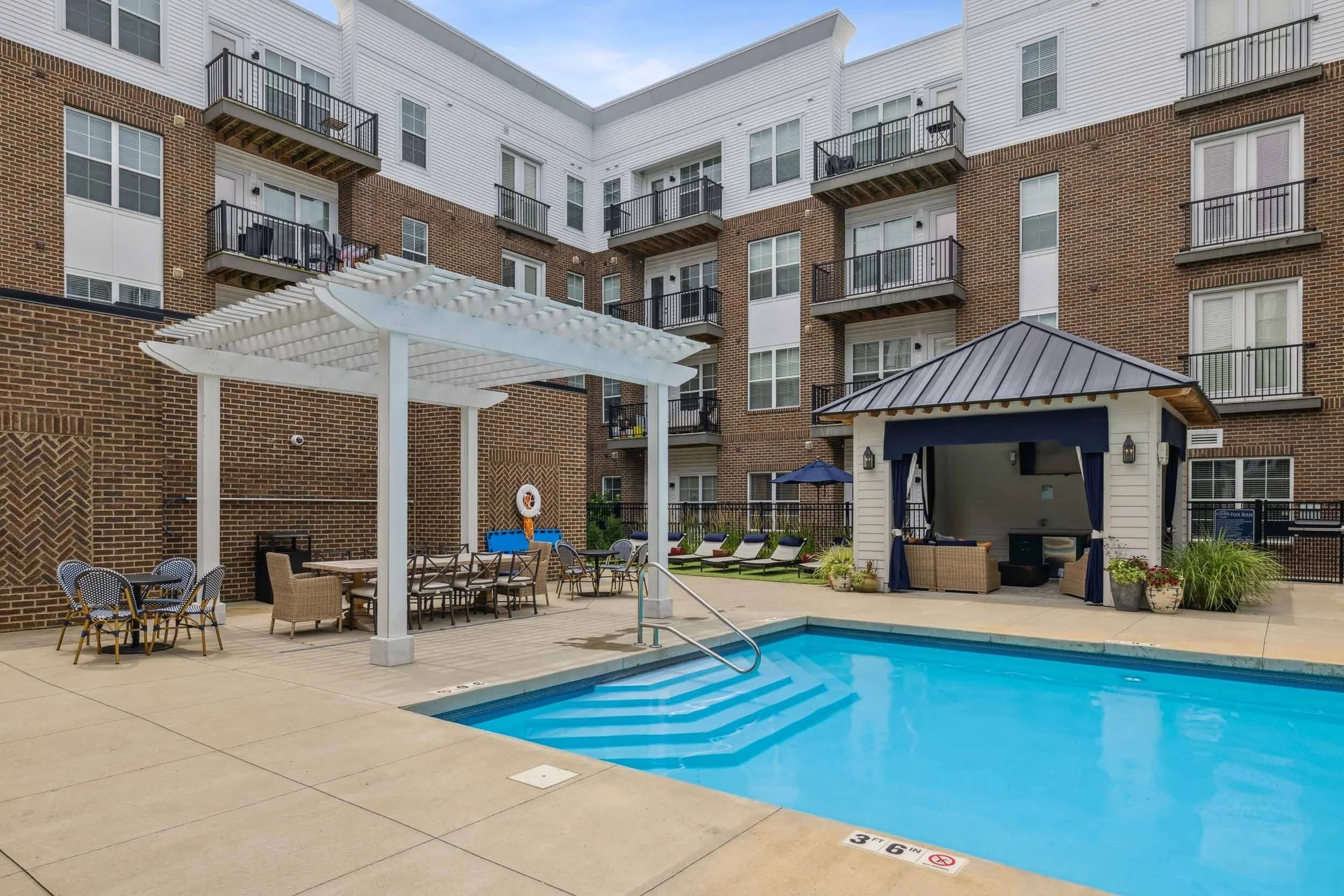 Outdoor pool area at an apartment complex with lounge chairs, pergola-covered seating, tables, and a pavilion in a courtyard surrounded by four-story buildings.