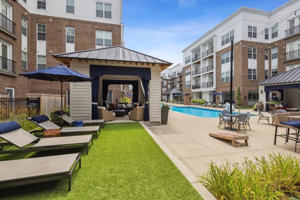 Outdoor pool area at a modern apartment complex featuring lounge chairs, shaded seating cabanas, and manicured landscaping on a sunny day.