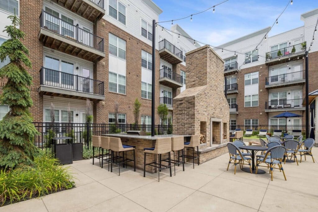 Outdoor patio area of a modern apartment complex with a brick fireplace, bar seating, patio tables, string lights, and surrounding greenery.