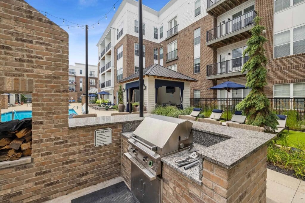 Outdoor grill station with a stainless steel barbecue in a courtyard area, surrounded by apartment buildings, lounge chairs, umbrellas, and a pool.