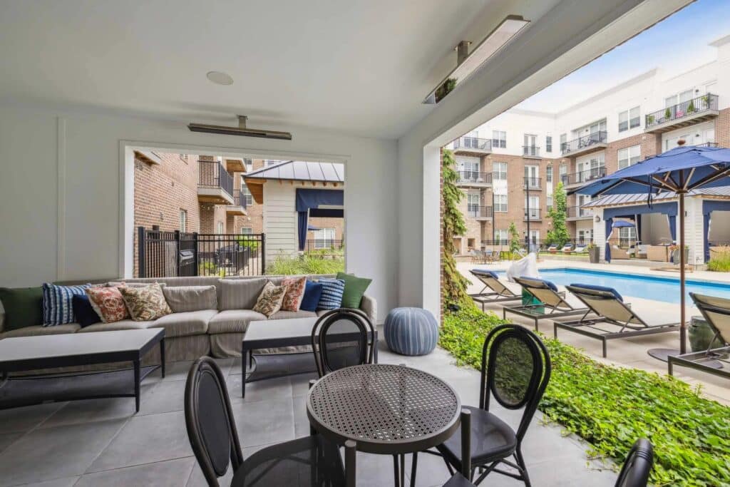 Covered patio area with a sofa and round table overlooks a swimming pool surrounded by lounge chairs and apartment buildings.