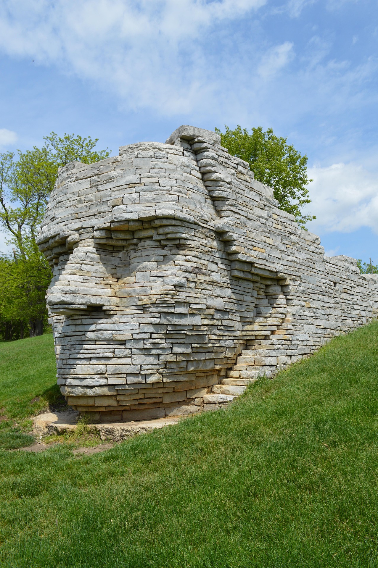 A large stone sculpture of a human face, constructed from stacked flat rocks, sits on a grassy hillside with trees and blue sky in the background.